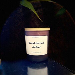 Pu&ograve; includere: Una candela di soia viola con un coperchio dorato e un'etichetta bianca che recita "Apothecary Sandalwood Amber Luxury Soy Candle".