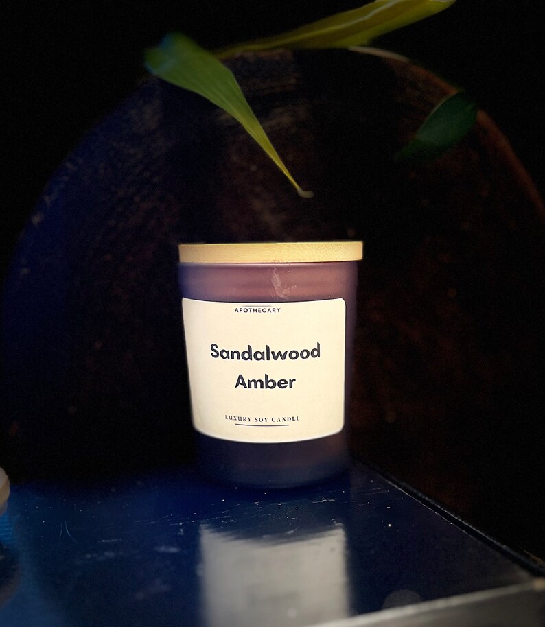 Pu&ograve; includere: Una candela di soia viola con un coperchio dorato e un'etichetta bianca che recita "Apothecary Sandalwood Amber Luxury Soy Candle".