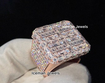 May include: A square-shaped ring with a rose gold band and a large, square-shaped setting covered in baguette-cut diamonds. The ring is held in a gloved hand. The text "Iceman Jewels" is visible on the ring and in the background.