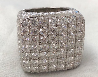 May include: A square-shaped ring with a silver band and a large, square-shaped diamond set in the center. The diamond is surrounded by smaller diamonds that are set in a pave setting.