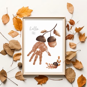 Op de afbeelding: Een ingelijste print met een herfstthema. Het kunstwerk toont een handafdruk, eikels en de woorden "hello FALL" in handschriftstijl. De print is omgeven door herfstbladeren in bruin- en oranjetinten.