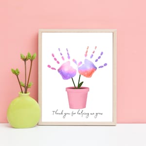 Op de afbeelding: Een ingelijst print met een handafdruk bloemenontwerp. De bloemen zijn roze, paars en oranje. De tekst luidt "Bedankt voor het helpen met groeien".