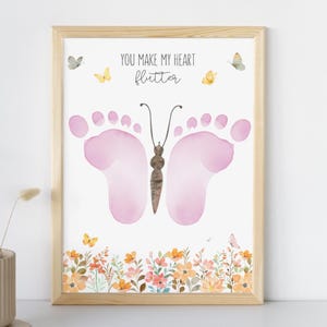 May include: A framed print with a butterfly design made of pink footprints. The text "YOU MAKE MY HEART flutter" is above the butterfly. The bottom of the print has a floral border with orange, pink, and yellow flowers. Butterflies are scattered around the design.