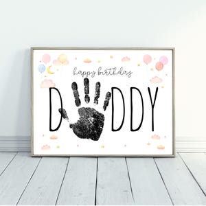 Pu&ograve; includere: Stampa incorniciata con le parole "happy birthday DADDY" in nero. Un'impronta di mano nera &egrave; al centro della parola "DADDY". La stampa &egrave; decorata con palloncini, nuvole e stelle ad acquerello in colori pastello.