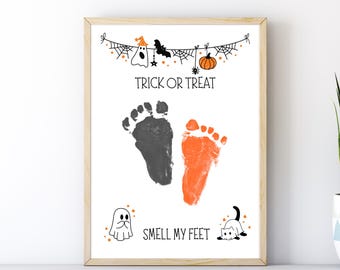 May include: A framed Halloween-themed print with a light wood frame. The print features two baby footprints, one black and one orange, with the text "TRICK OR TREAT" and "SMELL MY FEET". Decorative elements include a ghost, bat, and pumpkin.