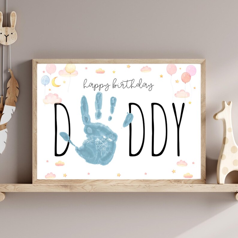 Pu&ograve; includere: Una stampa incorniciata con le parole "happy birthday DADDY" in corsivo nero e lettere maiuscole. Un'impronta di mano blu &egrave; al centro. Lo sfondo presenta palloncini, nuvole e stelle ad acquerello. La cornice &egrave; marrone chiaro.