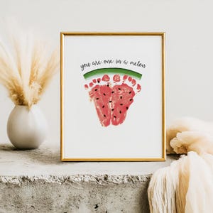 Peut inclure: Impression encadrée avec un cadre doré. Elle présente un motif de pastèque composé d'empreintes de pieds rouges avec des détails de graines noires. Le texte "you are one in a melon" est écrit au-dessus. L'impression est posée sur une surface en béton.