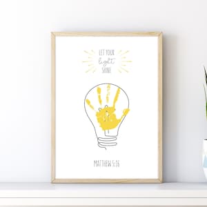 Puede incluir: Impresión artística enmarcada con una huella de mano amarilla dentro de un contorno de bombilla. Las palabras "LET YOUR light SHINE" están encima de la bombilla, y "MATTHEW 5:16" debajo. La obra de arte está sobre un fondo blanco y enmarcada en madera clara.