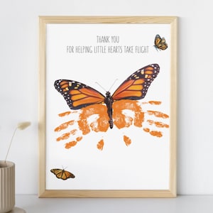May include: Framed print with a monarch butterfly illustration and orange handprints. The text reads, "THANK YOU FOR HELPING LITTLE HEARTS TAKE FLIGHT." The butterfly has orange and black wings. The frame is made of light-colored wood.