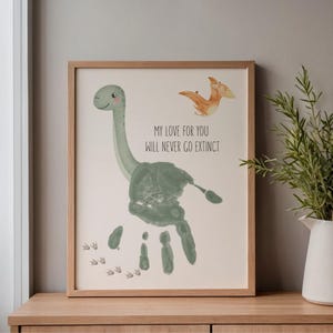 Op de afbeelding: Een ingelijste print met een handafdruk-dinosaurusontwerp in groentinten. Het kunstwerk toont een lachende Brontosaurus, een Pterodactylus en de tekst "MY LOVE FOR YOU WILL NEVER GO EXTINCT". Het frame is gemaakt van lichtgekleurd hout.