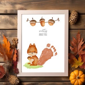 Op de afbeelding: Wit ingelijste print met een cartoon eekhoorn die een eikel vasthoudt, een voetafdruk en de tekst "nuts ABOUT YOU". De print heeft een herfstthema met eikels en een groene grasvlek.