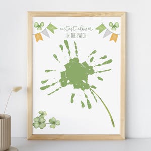 Può includere: Stampa incorniciata con cornice in legno chiaro. La stampa presenta un disegno di impronte di mani verdi che ricorda un dente di leone, con la scritta "cutest clover IN THE PATCH". Trifogli e banner ad acquerello completano il tema del giorno di San Patrizio.