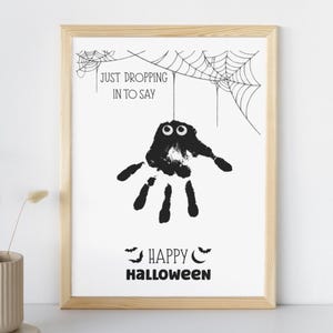 Puede incluir: Un estampado enmarcado con temática de Halloween con un diseño de araña de huella de mano. El estampado incluye el texto "JUST DROPPING IN TO SAY" encima de la araña y "HAPPY HALLOWEEN" debajo. El marco es de madera clara.