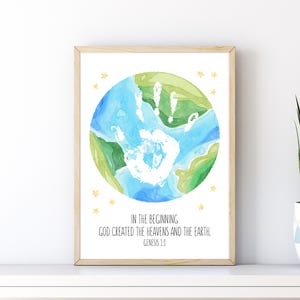 May include: Watercolor print in a light wood frame. The artwork depicts a blue and green globe with a white handprint in the center. The text reads "IN THE BEGINNING GOD CREATED THE HEAVENS AND THE EARTH. GENESIS 1:1". Yellow stars surround the globe.