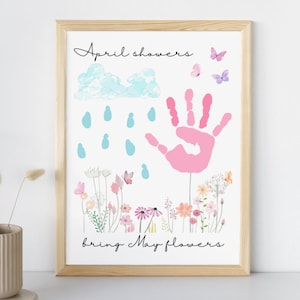 May include: A framed print with the words "April showers bring May flowers." The artwork features a pink handprint, blue raindrops, a cloud, butterflies, and colorful flowers. The frame is made of light-colored wood.
