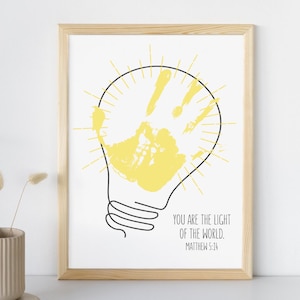 May include: A framed print featuring a yellow handprint inside a black outline of a lightbulb. The text "YOU ARE THE LIGHT OF THE WORLD. MATTHEW 5:14" is printed in black below the lightbulb. The frame is made of light-colored wood.