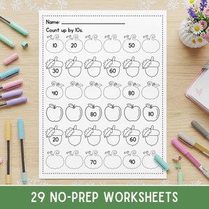 May include: A black and white worksheet with pumpkin, acorn, and apple illustrations for counting by tens. The worksheet includes the text "Count up by 10s" and "29 NO-PREP WORKSHEETS". Pens and a small bouquet of flowers are in the background.