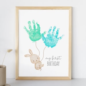 Puede incluir: Impresión enmarcada con dos huellas de manos en verde azulado y turquesa, que parecen globos, con un conejito beige sujetando las cuerdas. El texto "my first BIRTHDAY" está escrito en negro. El marco es de madera clara.