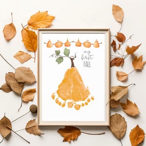 May include: A framed print featuring a pumpkin design made from a baby's footprint. The pumpkin is orange, with a brown stem and green leaves. The words "my first FALL" are written in black script. The print is surrounded by autumn leaves.