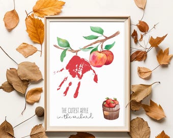 May include: A framed print featuring a red handprint shaped like an apple, a watercolor branch with two apples, and a small wooden basket filled with red apples. The text reads, The cutest apple in the orchard, in a simple black script font.