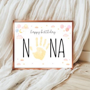May include: A framed print with a white background, featuring the words "happy birthday" in cursive above the word "NANA" in bold black letters. A yellow handprint is in the center. Pastel balloons, clouds, and stars decorate the top and bottom.