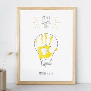 Puede incluir: Una impresión enmarcada con un marco de madera clara. La obra de arte presenta un contorno negro de una bombilla con una huella de mano amarilla en el interior. Encima de la bombilla está el texto "LET YOUR light SHINE" y debajo "MATTHEW 5:16".
