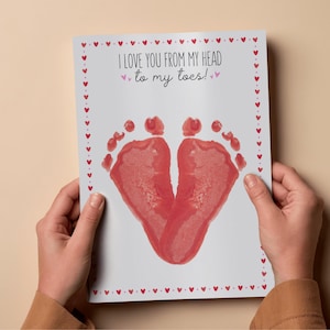 May include: A white rectangular greeting card featuring a border of small red hearts. The text reads I LOVE YOU FROM MY HEAD to my toes! in black lettering. Below the text are two red baby footprints positioned to form a heart shape on the card.