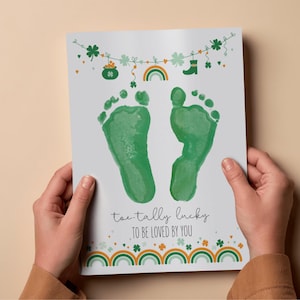 May include: A St. Patrick's Day themed print with two green baby footprints. The print features a garland of shamrocks, a pot of gold, a rainbow, and a green boot. The text reads "toe-tally lucky TO BE LOVED BY YOU."