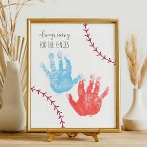 Può includere: Una stampa incorniciata con una cornice dorata. Presenta due impronte di mani, una blu e una rossa, su sfondo bianco. Il testo "always swing for the fences" è scritto in corsivo nero. Dettagli di cucitura rossi assomigliano a una palla da baseball.