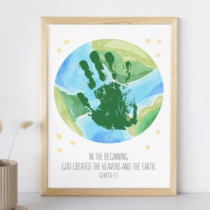 Può includere: Stampa artistica incorniciata con una rappresentazione ad acquerello della Terra, un'impronta di mano verde e stelle dorate. Il testo recita: "IN PRINCIPIO DIO CREÒ I CIELI E LA TERRA. GENESI 1:1". Cornice in legno chiaro.