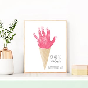 May include: A framed print featuring a pink handprint shaped like ice cream on a cone. The text reads "YOU ARE THE coolest HAPPY FATHER'S DAY!" The artwork is displayed on a white surface.