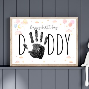 Pu&ograve; includere: Stampa incorniciata con le parole "happy birthday DADDY" e un'impronta di mano nera. La stampa &egrave; decorata con palloncini ad acquerello, nuvole e stelle dai colori pastello. La cornice &egrave; di colore legno chiaro.