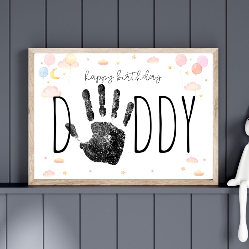 Pu&ograve; includere: Stampa incorniciata con le parole "happy birthday DADDY" e un'impronta di mano nera. La stampa &egrave; decorata con palloncini ad acquerello, nuvole e stelle dai colori pastello. La cornice &egrave; di colore legno chiaro.