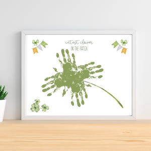 May include: A framed print with a green handprint clover design and the text "cutest clover in the patch." The artwork includes watercolor-style shamrocks and banner accents in orange, green, and grey. The print is displayed on a wooden surface.
