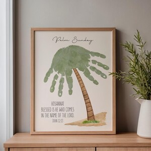 May include: Framed artwork with a palm tree design created from a handprint. The print includes the text "Palm Sunday" and a Bible verse. The frame is light brown, and the background is off-white. The artwork is displayed on a wooden surface.