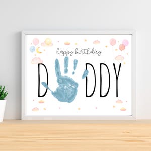 Pu&ograve; includere: Una stampa incorniciata con le parole "Happy Birthday Daddy" in corsivo nero. La parola "DADDY" &egrave; in grandi lettere nere con un'impronta blu al centro. Palloncini, nuvole e stelle pastello decorano la parte superiore.