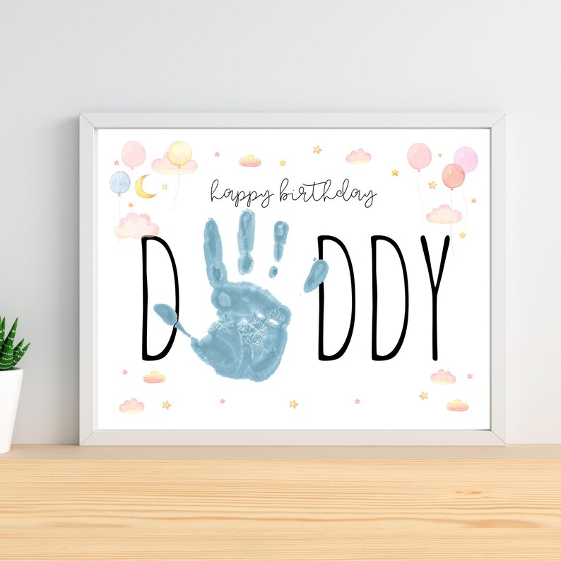 Pu&ograve; includere: Una stampa incorniciata con le parole "Happy Birthday Daddy" in corsivo nero. La parola "DADDY" &egrave; in grandi lettere nere con un'impronta blu al centro. Palloncini, nuvole e stelle pastello decorano la parte superiore.