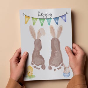May include: A watercolor print with the text "hoppy Easter" and a banner. The print features two bunny footprints, a chick, and a bunny in an egg. The banner has the letters "E A S T E R" on it.