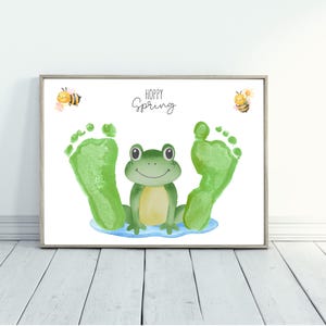 Puede incluir: Impresión enmarcada con una rana verde, huellas verdes y dos abejas de dibujos animados. Las palabras "Hoppy Spring" están escritas encima de la rana. La rana está sentada en un charco azul.
