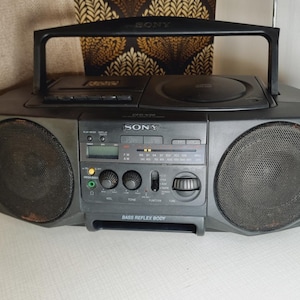 Puede incluir: Un reproductor de radio y casetes Sony negro con una cara plateada y dos altavoces grandes. La radio tiene una pantalla digital y una variedad de botones para controlar la radio y el reproductor de casetes. El texto "RADIO REFLEX BODY" está impreso en la parte frontal de la radio.