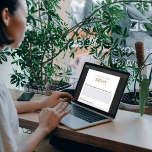 Op de afbeelding: Een persoon gebruikt een laptop met een enquêteformulier op het scherm. De laptop staat op een houten oppervlak, met een smartphone in de buurt. Groene planten zijn op de achtergrond te zien. De enquête heet "Client Base Lesson Survey".