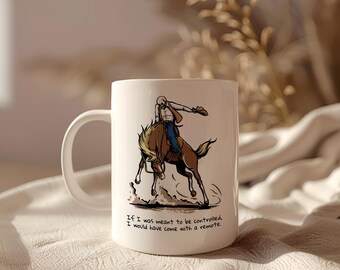 May include: White ceramic mug with a cartoon illustration of a person riding a bucking horse. The text on the mug reads "If I was meant to be controlled, I would have come with a remote."