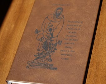 May include: A brown leather-bound journal with a religious illustration and text. The image features a figure pouring water over a skeleton. The text reads "Therefore, if anyone is in Christ, he is a new creation. The old has passed away; behold, the new has come."