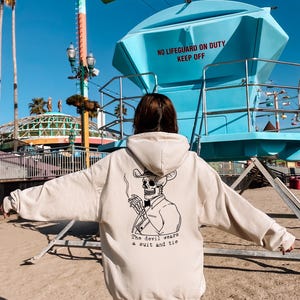 May include: Beige hoodie with a skeleton wearing a cowboy hat and smoking a cigarette. The text "The devil wears a suit and tie" is below the graphic. The background includes a lifeguard station and amusement park rides.