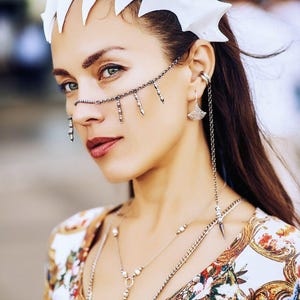 May include: A woman wearing a white crown with a silver chain nose ring and multiple silver necklaces. She is wearing a white and gold patterned blouse.
