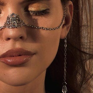 May include: A woman wearing a silver nose chain with a decorative floral design. The chain extends to a dangling crystal and tassel earring.
