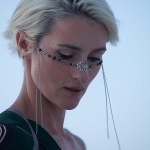 Puede incluir: Una mujer con el pelo corto y rubio platino que lleva un aro de nariz de cadena plateada y una máscara facial de cadena plateada con cuentas negras. La máscara está unida a las orejas con cadenas plateadas. Lleva una camisa con estampado verde y negro.