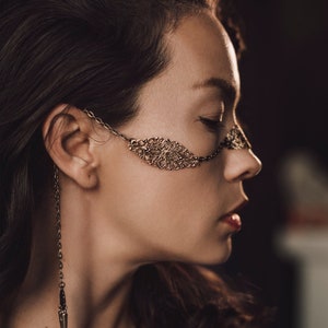 May include: A woman wearing a delicate gold chain face mask with a detailed floral design. The chain is attached to her ear and extends to the other side of her face. She is also wearing a gold chain necklace with a pendant.