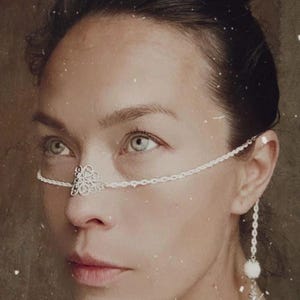 May include: A woman wearing a delicate silver nose chain with a floral design. The chain is attached to a small silver earring with a pearl accent.