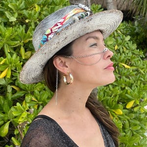 May include: A woman wearing a large, white, weathered straw hat with a colorful patterned band. She is also wearing a silver chain nose ring and large gold hoop earrings.